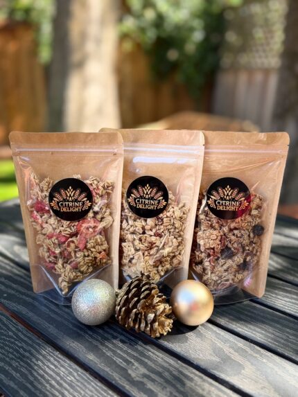 Citrine Delight Strawberry Almond, Coconut and Pecan Berry granola pouches in a row with holiday ornaments on a wood table.